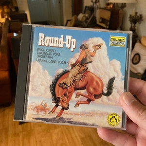May include: A CD case with a blue background and a cowboy riding a bucking horse. The text on the case reads "Round-Up" and "Erich Kunzel Cincinnati Pops Orchestra Frankie Laine, Vocals".
