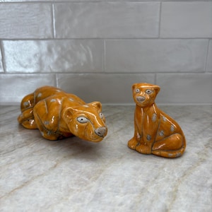 May include: Two ceramic leopard figurines. One is lying down, the other is sitting upright. Both are orange with white and gray spots, and have detailed facial features.