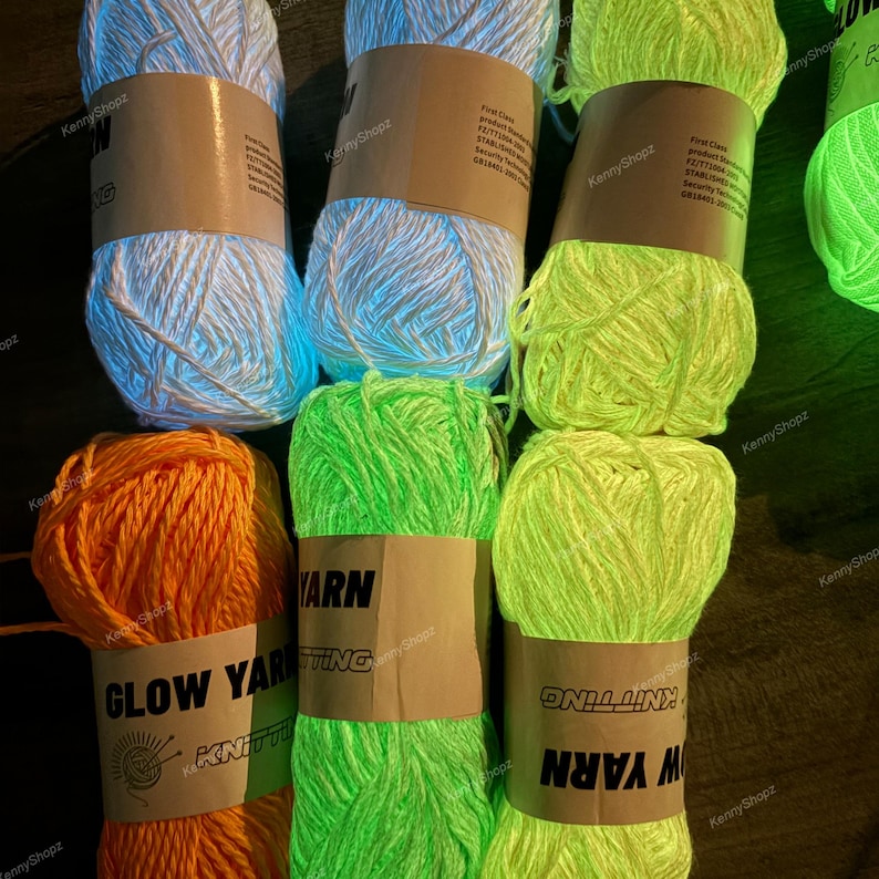 May include: Six skeins of glow-in-the-dark yarn arranged in two rows. The top row features white, light blue, and neon green yarn. The bottom row features orange, bright green, and yellow-green yarn. Each skein has a tan paper label.