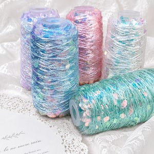 May include: Five spools of decorative thread in shades of blue, pink, and purple, wrapped with iridescent sequins. One spool lies on its side. A white card with the text "be no more rain" is visible.
