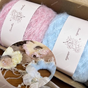 May include: Two skeins of fuzzy yarn in pastel pink and light blue, each wrapped in a paper band labeled Camellia studio with a floral illustration. Below, three circular hair accessories feature ruffled edges, lace, pearls, and beads.