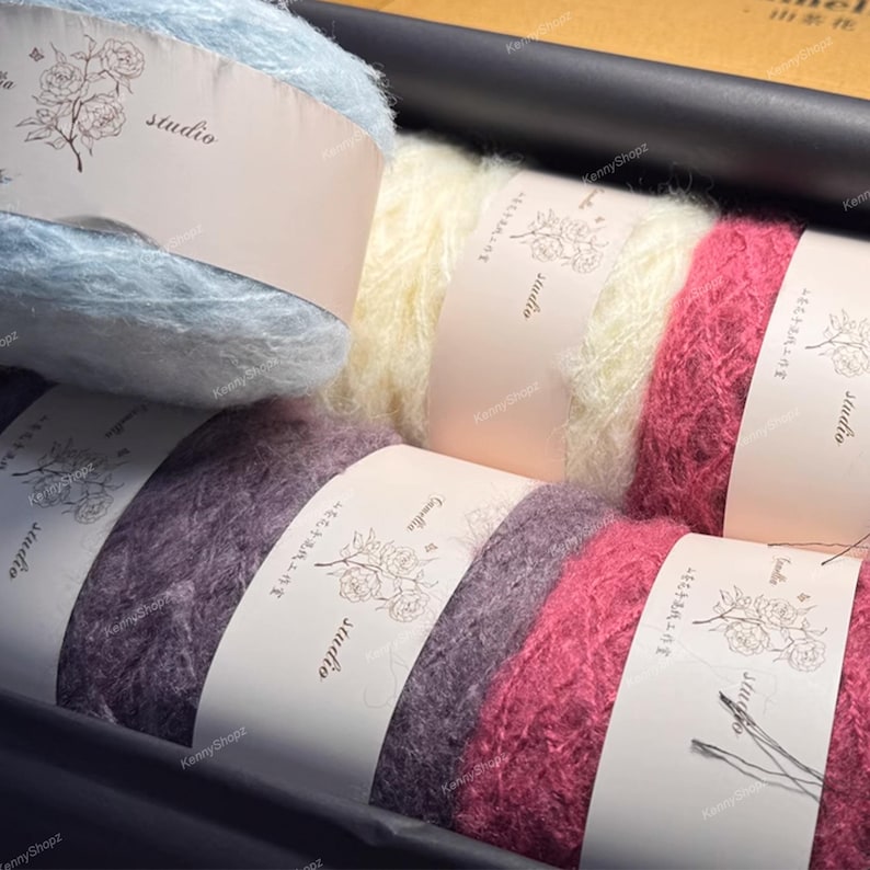 May include: A collection of six skeins of fuzzy yarn in light blue, cream, dark pink, purple, magenta, and deep red. Each skein is wrapped in a light beige paper band featuring a floral line drawing and the word studio printed in black text.