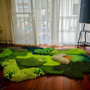 May include: A large, irregularly shaped rug with a landscape design. The rug features various shades of green, with textured areas resembling grass, hills, and small pom-pom accents. The rug is placed on a dark wood floor.