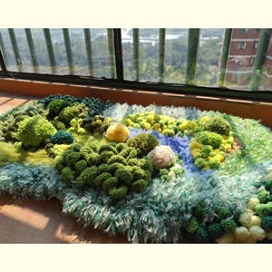 May include: A textured rug featuring a landscape design with various shades of green, yellow, and blue yarn. The surface is composed of tufted, pom-pom, and shaggy elements that create a three-dimensional, moss-like appearance across the mat.