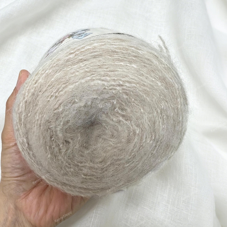 May include: A round ball of light beige, fuzzy yarn with a soft, textured appearance. The yarn features a subtle heathered effect with lighter and darker fibers blended throughout. A paper label is partially visible on the top of the yarn ball.