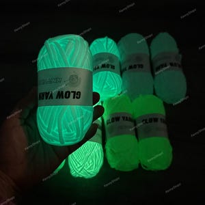 May include: Several skeins of glow-in-the-dark yarn emitting a bright neon green light in a dark environment. Each skein features a white paper label with the text GLOW YARN and a small knitting icon. The yarn has a thick, soft texture.