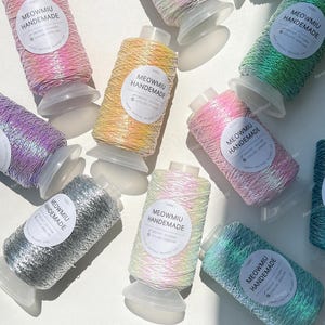 May include: Several spools of iridescent yarn in various colours, including pink, yellow, and green. Each spool has a label reading "MEOWMIU HANDEMADE." The yarn is wound onto a plastic base. The image is taken from above, showing the spools scattered.