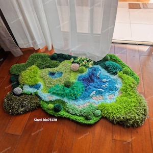 K&ouml;nnte beinhalten: Ein getufteter Teppich mit unregelm&auml;&szlig;iger Form, der eine Landschaft mit gr&uuml;nen H&uuml;geln, einem blauen See und kleinen texturierten Pompons zeigt. Der Teppich weist verschiedene Gr&uuml;n- und Blaut&ouml;ne auf und tr&auml;gt die Gr&ouml;&szlig;enangabe 130x75CM.