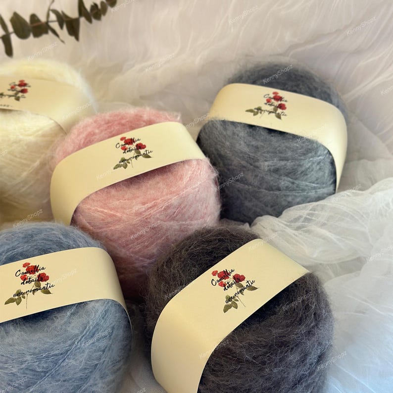 May include: Five round balls of fuzzy yarn in cream, light pink, dark gray, light blue, and charcoal gray. Each ball features a cream-colored paper band with a printed illustration of a red rose and the text Camellia details aeromantic.