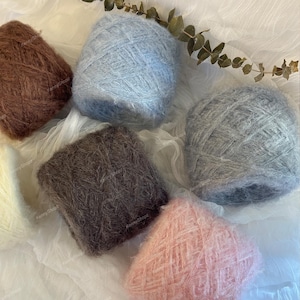 May include: A collection of six round balls of fuzzy yarn in various colors including brown, light blue, dark gray, light gray, and soft pink, arranged together. Each ball of yarn features a soft, textured, and slightly hairy appearance.