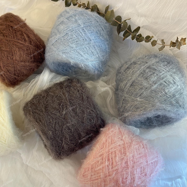 May include: A collection of six round balls of fuzzy yarn in various colors including brown, light blue, dark gray, light gray, and soft pink, arranged together. Each ball of yarn features a soft, textured, and slightly hairy appearance.