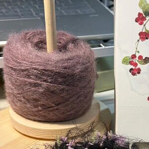 May include: A dusty mauve yarn cake mounted on a light-colored wooden yarn holder with a vertical spindle. The yarn has a fuzzy, textured appearance. A portion of a white card featuring a botanical illustration of red berries is visible nearby.