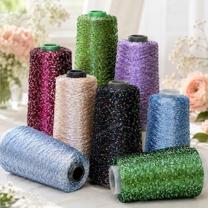 May include: A collection of eight conical spools of metallic thread in various colours including deep red, bright green, light purple, beige, black with multicoloured flecks, and two shades of blue, arranged in a cluster on a flat surface.