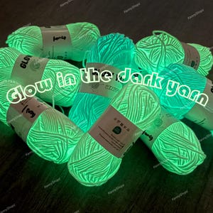 Puede incluir: Una colección de varias madejas de hilo que brillan en la oscuridad con una luz verde neón brillante. Cada madeja está envuelta en una etiqueta de papel con el texto Glow in the dark yarn, junto con varios símbolos de cuidado de lavado.