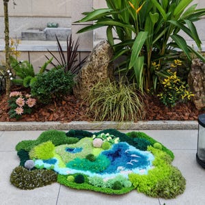 K&ouml;nnte beinhalten: Ein getufteter Teppich mit einem abstrakten Landschaftsmotiv aus verschiedenfarbigen gr&uuml;nen, blauen und wei&szlig;en Garnen. Der Teppich hat eine moosartige Textur mit erhabenen, kreisf&ouml;rmigen B&uuml;scheln und einem geschwungenen blauen Flussmuster.