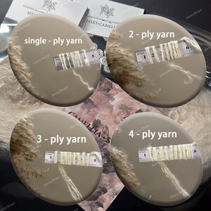 May include: Four circular beige displays showing yarn samples labeled single-ply, 2-ply, 3-ply, and 4-ply yarn. Each display features a metal ruler measuring the thickness of the cream-colored yarn strands in centimeters, with visible text markings.