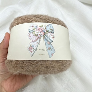 May include: A ball of fuzzy, light brown yarn wrapped in a cream-colored paper band. The band features a central illustration of a large bow decorated with pink roses and blue floral patterns, along with the text Camellia.