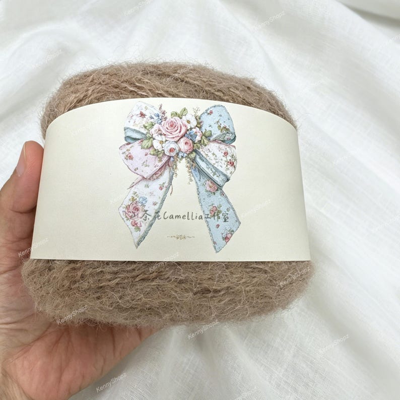 May include: A ball of fuzzy, light brown yarn wrapped in a cream-colored paper band. The band features a central illustration of a large bow decorated with pink roses and blue floral patterns, along with the text Camellia.
