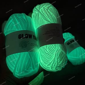 May include: Three skeins of yarn that glow with a bright neon green light in the dark. Each skein features a white paper label with the text GLOW YARN and a graphic of a ball of yarn with knitting needles. The yarn has a thick, textured appearance.