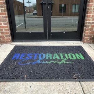a mat with the text "Restoration Church" printed on it, placed in front of a glass door.