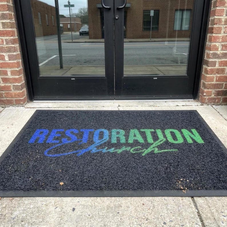 a mat with the text "Restoration Church" printed on it, placed in front of a glass door.