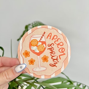 K&ouml;nnte beinhalten: Runder Untersetzer mit einem pfirsichfarbenen und wei&szlig;en gestreiften Rand. Der Untersetzer zeigt eine Grafik eines Aperol Spritz Cocktails mit Orangenscheiben und den Worten "Aperol Spritz". Orangefarbene Blumenillustrationen sind ebenfalls enthalten.