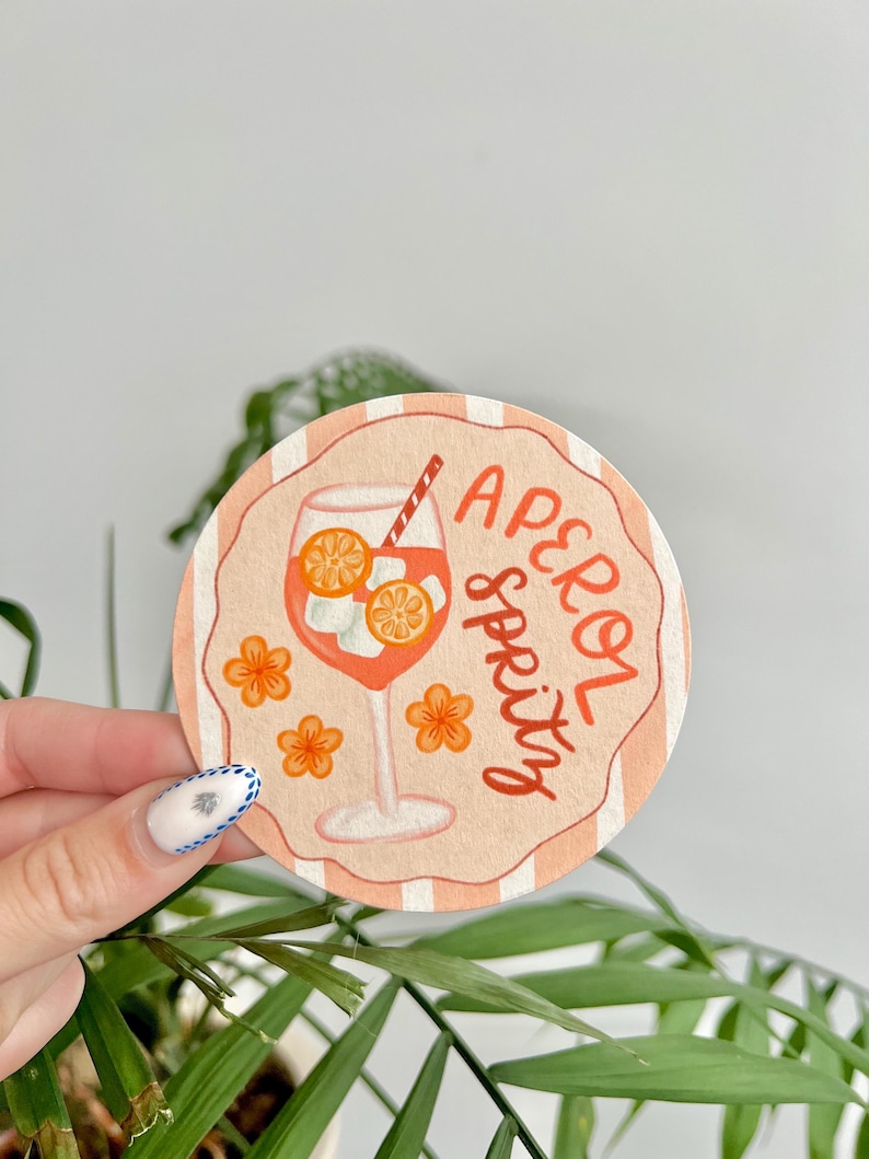 K&ouml;nnte beinhalten: Runder Untersetzer mit einem pfirsichfarbenen und wei&szlig;en gestreiften Rand. Der Untersetzer zeigt eine Grafik eines Aperol Spritz Cocktails mit Orangenscheiben und den Worten "Aperol Spritz". Orangefarbene Blumenillustrationen sind ebenfalls enthalten.