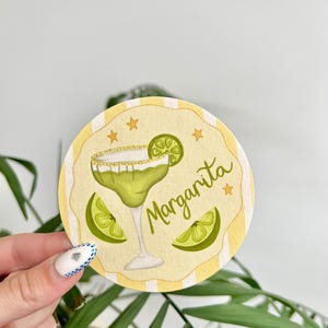 K&ouml;nnte beinhalten: Ein runder Untersetzer mit einer Margarita-Illustration. Der Untersetzer zeigt einen gr&uuml;nen Margarita-Cocktail mit einer Limettenscheibe und goldenem Rand sowie das Wort "Margarita". Der Hintergrund ist gelb mit vertikalen Streifen und kleinen goldenen Sternen.