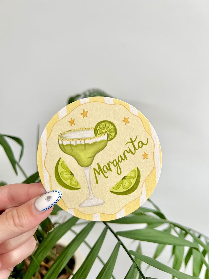 K&ouml;nnte beinhalten: Ein runder Untersetzer mit einer Margarita-Illustration. Der Untersetzer zeigt einen gr&uuml;nen Margarita-Cocktail mit einer Limettenscheibe und goldenem Rand sowie das Wort "Margarita". Der Hintergrund ist gelb mit vertikalen Streifen und kleinen goldenen Sternen.