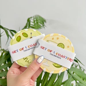 K&ouml;nnte beinhalten: Ein Set aus vier Untersetzern mit Cocktail-Motiv. Jeder Untersetzer zeigt eine andere Cocktail-Illustration, darunter Oliven und eine Limette. Die Untersetzer sind rund mit einem gelb-wei&szlig; gestreiften Hintergrund. Der Text "SET OF 4 COASTERS" ist auf einem wei&szlig;en Band gedruckt.