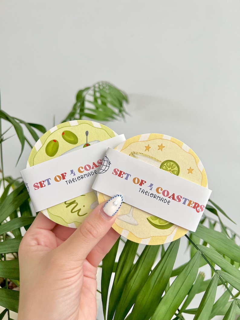 K&ouml;nnte beinhalten: Ein Set aus vier Untersetzern mit Cocktail-Motiv. Jeder Untersetzer zeigt eine andere Cocktail-Illustration, darunter Oliven und eine Limette. Die Untersetzer sind rund mit einem gelb-wei&szlig; gestreiften Hintergrund. Der Text "SET OF 4 COASTERS" ist auf einem wei&szlig;en Band gedruckt.