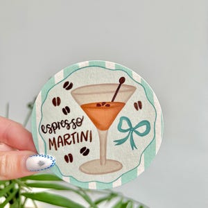 K&ouml;nnte beinhalten: Ein runder Untersetzer mit einem t&uuml;rkis-wei&szlig; gestreiften Rand. Der Untersetzer zeigt eine Illustration eines Espresso Martini mit Kaffeebohnen und einer t&uuml;rkisfarbenen Schleife. Die Worte "espresso MARTINI" sind geschrieben.
