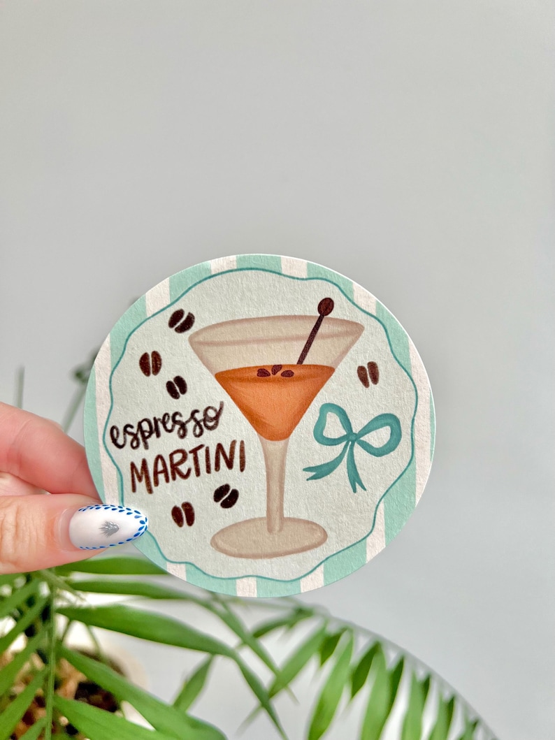 K&ouml;nnte beinhalten: Ein runder Untersetzer mit einem t&uuml;rkis-wei&szlig; gestreiften Rand. Der Untersetzer zeigt eine Illustration eines Espresso Martini mit Kaffeebohnen und einer t&uuml;rkisfarbenen Schleife. Die Worte "espresso MARTINI" sind geschrieben.