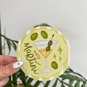 K&ouml;nnte beinhalten: Runder, hellgr&uuml;ner Untersetzer mit einer Aquarellillustration eines Dirty Martini. Das Martiniglas ist mit einer gelben Fl&uuml;ssigkeit gef&uuml;llt und mit Oliven garniert. Die Worte "Dirty Martini" sind auf dem Untersetzer geschrieben.