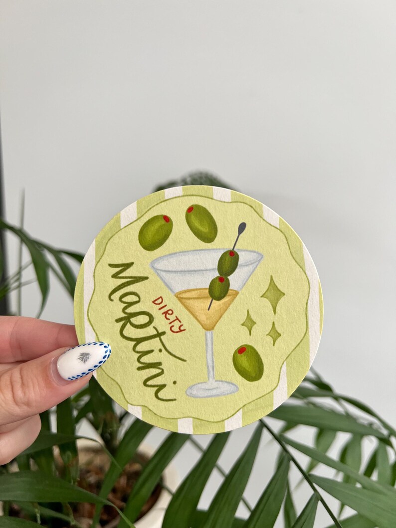 K&ouml;nnte beinhalten: Runder, hellgr&uuml;ner Untersetzer mit einer Aquarellillustration eines Dirty Martini. Das Martiniglas ist mit einer gelben Fl&uuml;ssigkeit gef&uuml;llt und mit Oliven garniert. Die Worte "Dirty Martini" sind auf dem Untersetzer geschrieben.
