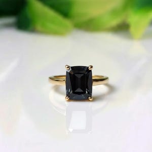May include: A gold ring with a large, rectangular, black gemstone. The ring has a simple band and four prongs holding the dark stone. The background is blurred, with green foliage and a white surface.