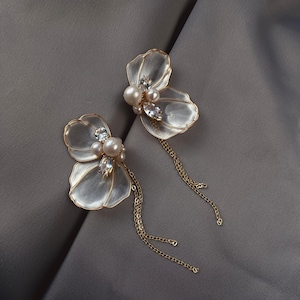 May include: A pair of earrings featuring translucent, petal-shaped elements with gold-toned wire edges. Each earring is accented with a cluster of white pearls and clear marquise-cut crystals, with a delicate gold-toned chain dangling from the base.