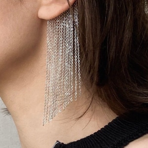 May include: A silver chain earring with a long, cascading fringe of smaller chains. The earring is worn in the ear of a person with dark hair.