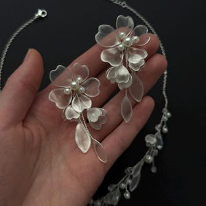K&ouml;nnte beinhalten: Ein silberfarbenes Blumen-Schmuckset mit Halskette und passenden Ohrringen. Die Ohrringe und die Halskette sind mit zarten, durchscheinenden Bl&uuml;tenbl&auml;ttern und Kunstperlen verziert. Der Schmuck wird vor dunklem Hintergrund pr&auml;sentiert.
