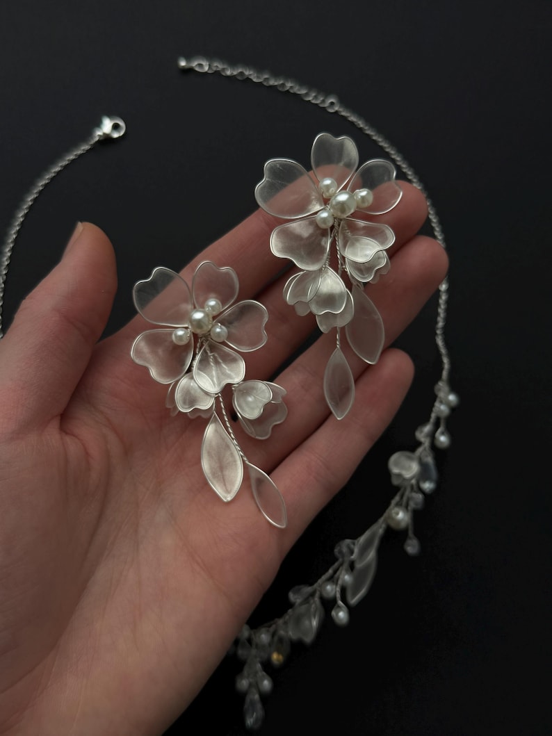 K&ouml;nnte beinhalten: Ein silberfarbenes Blumen-Schmuckset mit Halskette und passenden Ohrringen. Die Ohrringe und die Halskette sind mit zarten, durchscheinenden Bl&uuml;tenbl&auml;ttern und Kunstperlen verziert. Der Schmuck wird vor dunklem Hintergrund pr&auml;sentiert.