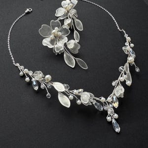 K&ouml;nnte beinhalten: Ein silberfarbenes Brautschmuck-Set mit floralem Design. Das Set umfasst eine Halskette und ein Haarteil, beide verziert mit klaren Kristallen, Kunstperlen und zarten Blumenakzenten. Der Schmuck ist vor einem schwarzen Hintergrund platziert.