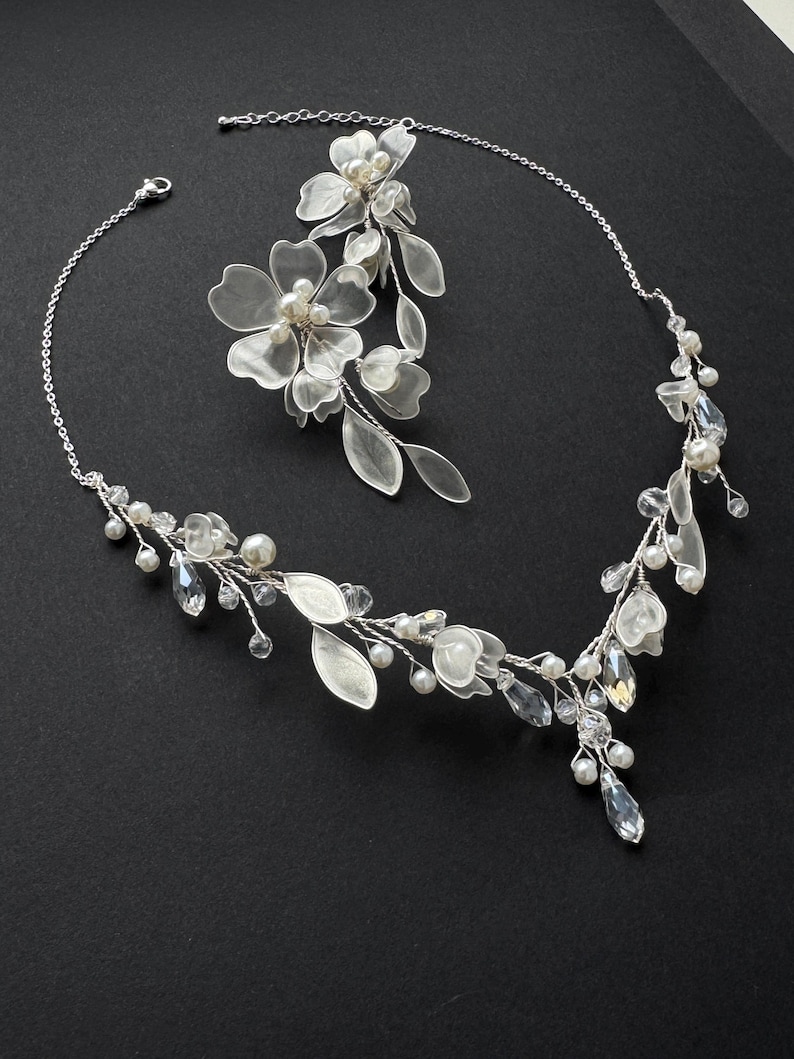 K&ouml;nnte beinhalten: Ein silberfarbenes Brautschmuck-Set mit floralem Design. Das Set umfasst eine Halskette und ein Haarteil, beide verziert mit klaren Kristallen, Kunstperlen und zarten Blumenakzenten. Der Schmuck ist vor einem schwarzen Hintergrund platziert.