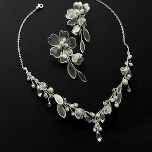 K&ouml;nnte beinhalten: Ein filigranes, silberfarbenes Brautschmuck-Set, bestehend aus einer Halskette und Ohrringen. Die Halskette und die Ohrringe sind mit durchscheinenden Bl&uuml;ten- und blattf&ouml;rmigen Akzenten, Perlen und kristallartigen Verzierungen versehen. Das Set wird vor schwarzem Hintergrund pr&auml;sentiert.