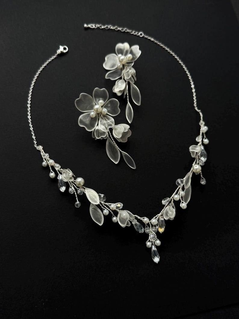 K&ouml;nnte beinhalten: Ein filigranes, silberfarbenes Brautschmuck-Set, bestehend aus einer Halskette und Ohrringen. Die Halskette und die Ohrringe sind mit durchscheinenden Bl&uuml;ten- und blattf&ouml;rmigen Akzenten, Perlen und kristallartigen Verzierungen versehen. Das Set wird vor schwarzem Hintergrund pr&auml;sentiert.