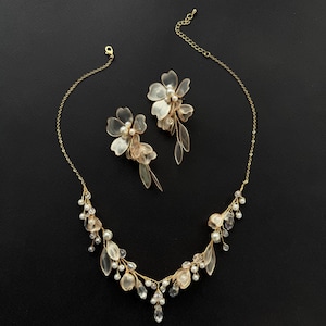 K&ouml;nnte beinhalten: Ein goldfarbenes Halsketten- und Ohrring-Set. Die Halskette und die Ohrringe weisen zarte, durchscheinende Blumen- und Blattmotive auf, die mit kleinen Perlen und klaren Kristallen akzentuiert sind. Der Schmuck ist vor einem schwarzen Hintergrund platziert.