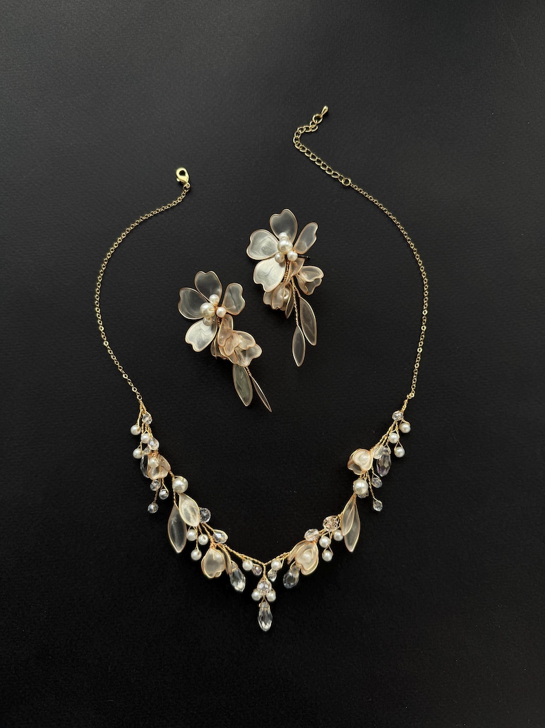 K&ouml;nnte beinhalten: Ein goldfarbenes Halsketten- und Ohrring-Set. Die Halskette und die Ohrringe weisen zarte, durchscheinende Blumen- und Blattmotive auf, die mit kleinen Perlen und klaren Kristallen akzentuiert sind. Der Schmuck ist vor einem schwarzen Hintergrund platziert.