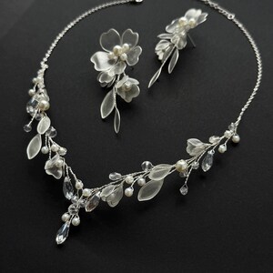 K&ouml;nnte beinhalten: Ein filigranes, silberfarbenes Brautschmuck-Set mit Halskette und passenden Ohrringen. Die Halskette und Ohrringe sind mit klaren Kristallen, Kunstperlen und durchscheinenden Bl&uuml;ten- und Blattakzenten verziert. Das Set wird vor schwarzem Hintergrund pr&auml;sentiert.