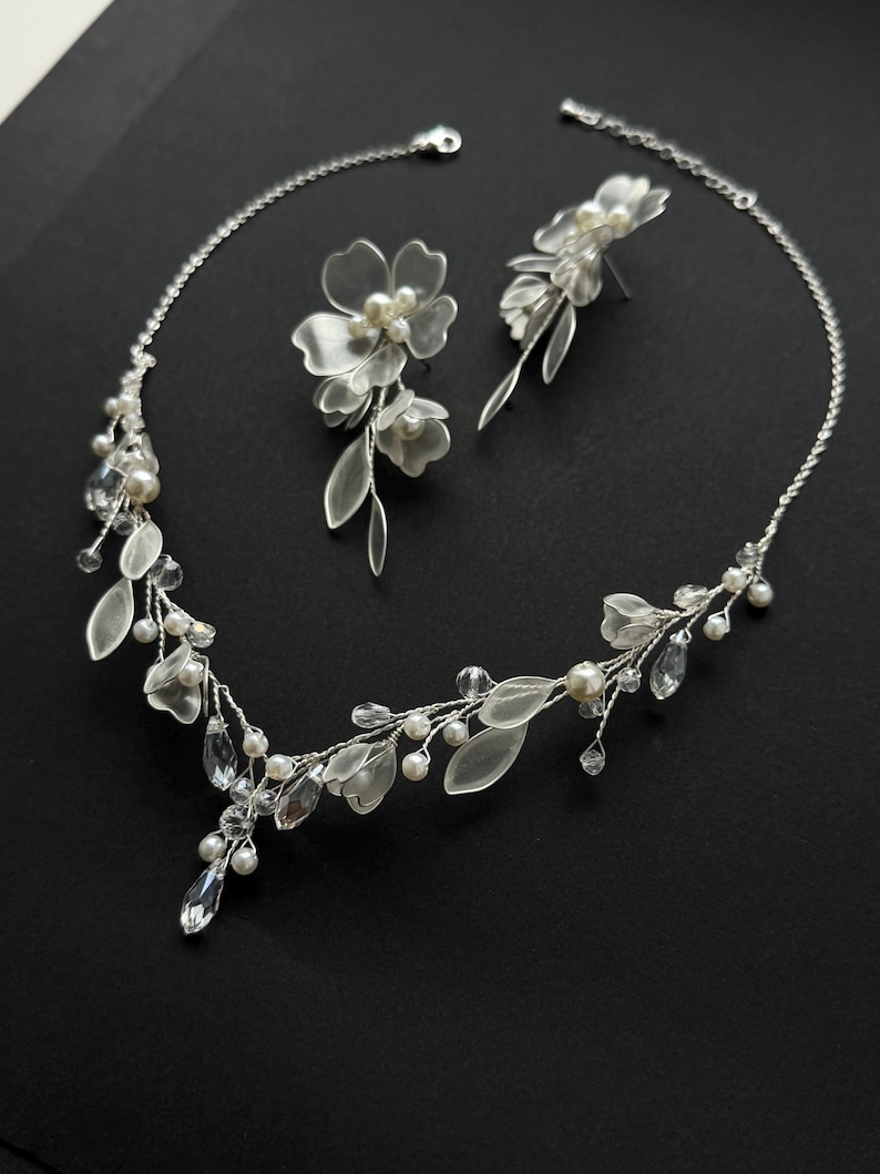 K&ouml;nnte beinhalten: Ein filigranes, silberfarbenes Brautschmuck-Set mit Halskette und passenden Ohrringen. Die Halskette und Ohrringe sind mit klaren Kristallen, Kunstperlen und durchscheinenden Bl&uuml;ten- und Blattakzenten verziert. Das Set wird vor schwarzem Hintergrund pr&auml;sentiert.
