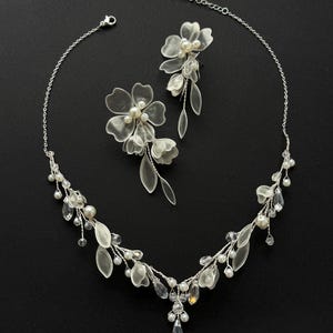 K&ouml;nnte beinhalten: Ein filigranes silberfarbenes Halsketten- und Ohrring-Set. Die Halskette und Ohrringe haben klare Kristalle, Kunstperlen und durchscheinende Bl&uuml;ten- und Bl&auml;tter-Akzente. Das Schmuckset wird vor einem schwarzen Hintergrund pr&auml;sentiert.