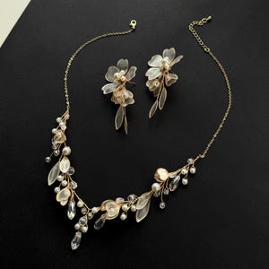 K&ouml;nnte beinhalten: Ein zartes, goldfarbenes Halsketten- und Ohrring-Set. Die Halskette und die Ohrringe verf&uuml;gen &uuml;ber florale und blattf&ouml;rmige Akzente mit Kunstperlen und klaren Kristallverzierungen. Der Schmuck wird auf einer schwarzen Oberfl&auml;che pr&auml;sentiert.